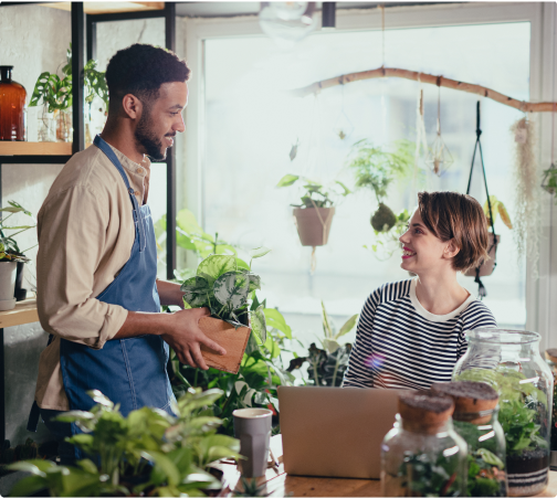 One man in a blue apron smiling towards a woman in a stripe long sleeve in a space with a lot of plants.